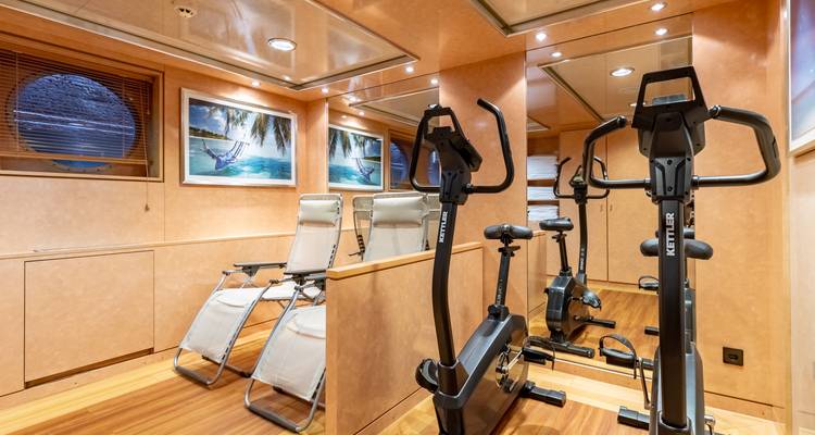 Kleiner Fitnessraum auf einem Kreuzfahrtschiff mit Heimtrainern, Liegen und maritimer Kunstwerk.