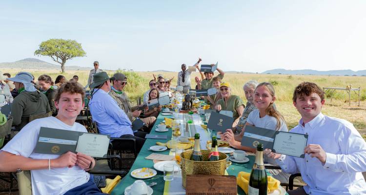 Gelukkige reizigers zittend aan een lange safaritafel houden trots vliegbewijzen omhoog in een open savannelandschap