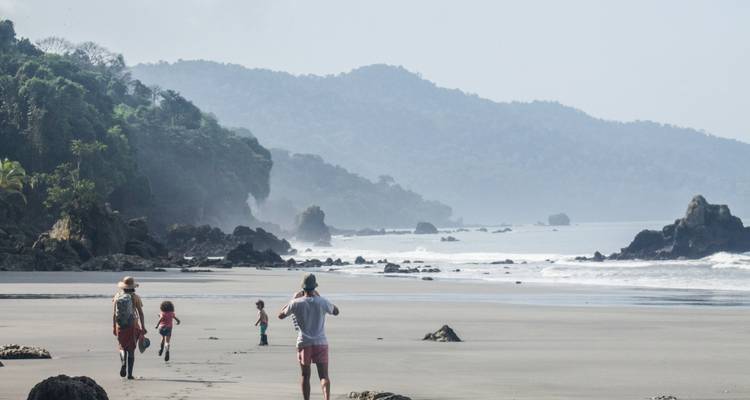 Familia caminando por una playa serena con acantilados rocosos en la distancia.