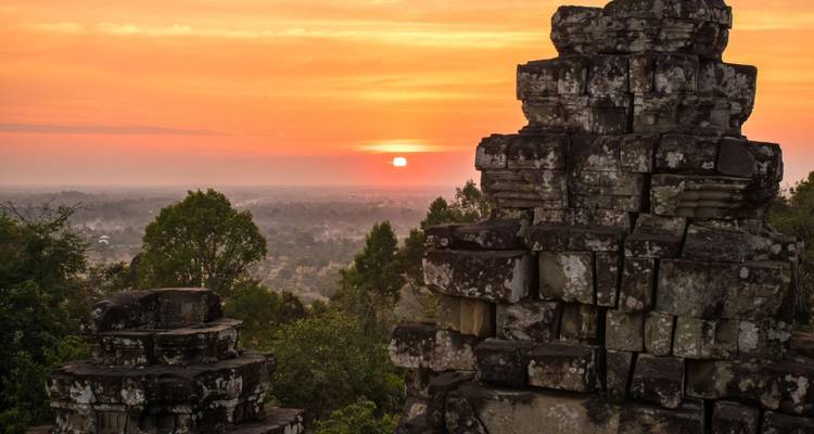 Alte Steintempelanlagen, die sich gegen einen leuchtend orangefarbenen Sonnenuntergang abheben, wobei die Sonne sich dem bewaldeten Horizont nähert.