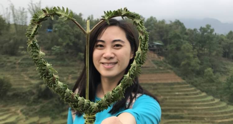 Frau, die eine herzförmige Blattstruktur hält, mit terrassierten Feldern im Hintergrund.