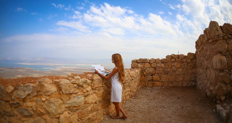 Persona leyendo mientras está de pie sobre un antiguo muro de piedra con vista al Mar Muerto.
