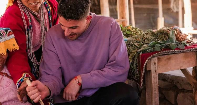 Traveler in a purple sweater learns a traditional craft from a local woman inside a rustic market stall filled with herbs.