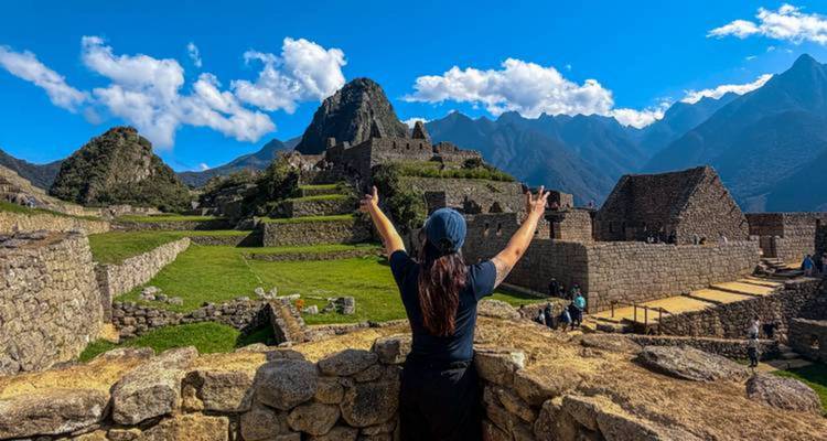 Fotografía de la mujer de espaldas levantando los brazos hacia la ciudadela de piedra de Machu Picchu bajo un cielo despejado.