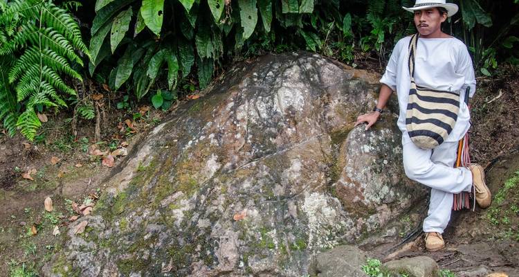Eine Person in traditioneller Kleidung, die sich in einer üppigen Umgebung an einen Felsen lehnt.