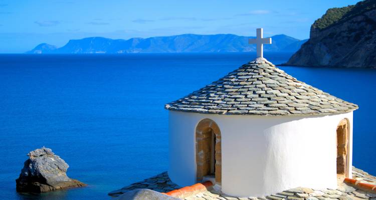 Pequeña capilla blanca con techo de piedra y cruz que domina el profundo mar Egeo azul.