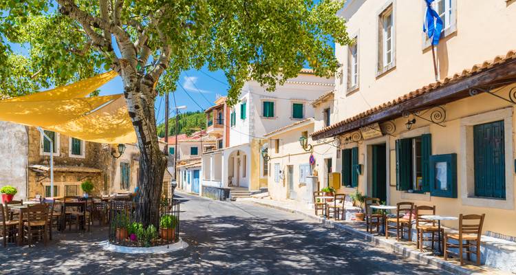 Calle soleada de un pueblito griego llena de cafés, casas encaladas y un plátano frondoso que da sombra.
