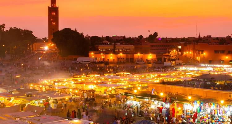 Un marché animé au coucher du soleil avec une haute tour de mosquée en arrière-plan.