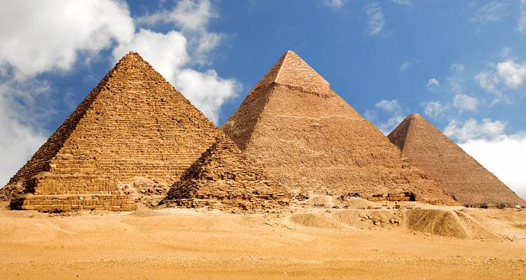 Three great pyramids of Giza dominate a sandy plateau under a partly cloudy sky.