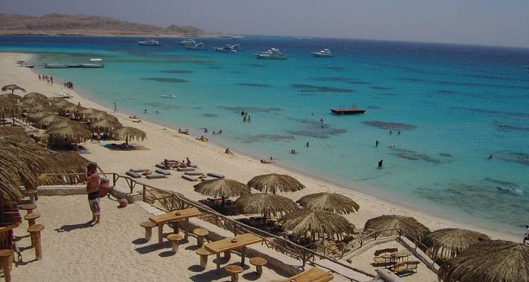 Playa tropical del Mar Rojo con agua turquesa, sombrillas de paja, tumbonas y bañistas, vista desde una terraza elevada.