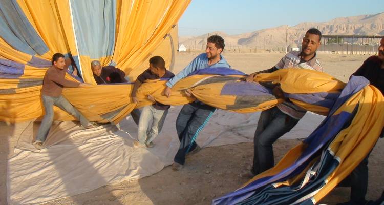 Los miembros de la tripulación luchan juguetonamente por extender la colorida envoltura de un globo aerostático en un campo de lanzamiento desértico.