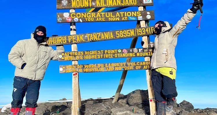 Two people near the Uhuru Peak sign, raising their hands with trekking poles.