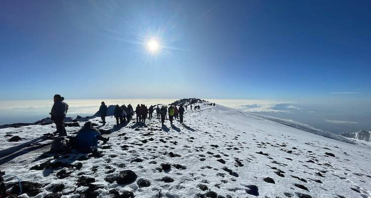 Group of people walking along a snowy trail on a mountain peak with the sun shining brightly.