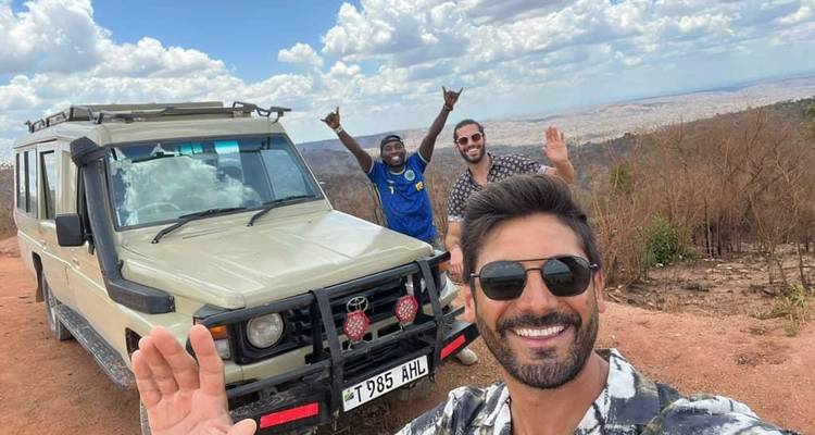 Tres turistas posando felizmente con un vehículo de safari y un fondo escénico.
