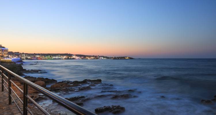 Küstenblick bei Sonnenuntergang mit ruhigem Meer und entfernten Gebäuden.
