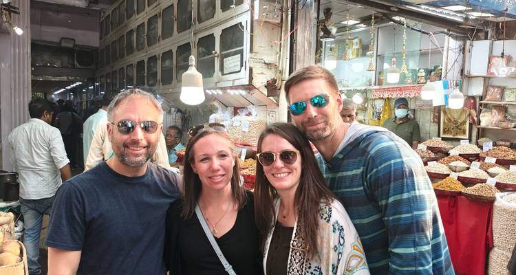 Grupo de cuatro turistas sonrientes parados juntos en un mercado de especias y frutos secos muy concurrido, con frascos y canastas apilados detrás de ellos.
