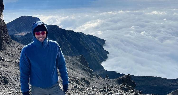 Eine Person in einer blauen Jacke, die auf einem felsigen Berggipfel über den Wolken steht.