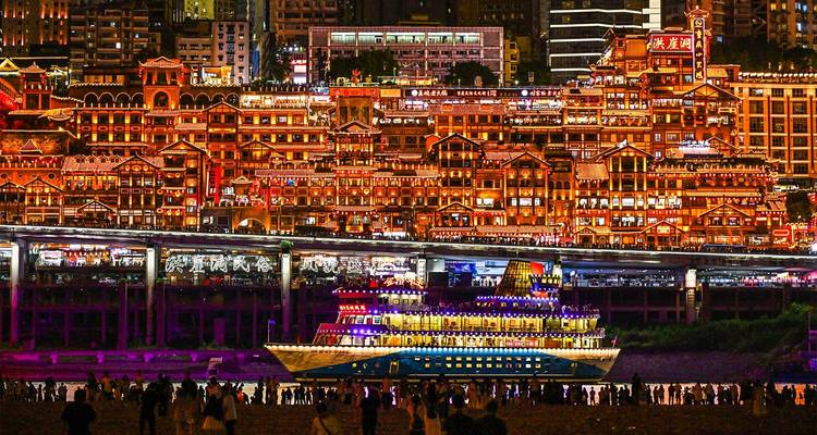 Vista nocturna de los edificios sobre pilotes escalonados de Hongyadong iluminados con luces naranjas sobre un crucero brillantemente iluminado en Chongqing.