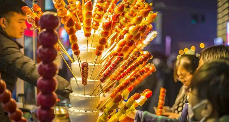 Vendeur du marché nocturne vendant des rangées de brochettes de fruits confits brillants illuminées par des lumières chaudes.