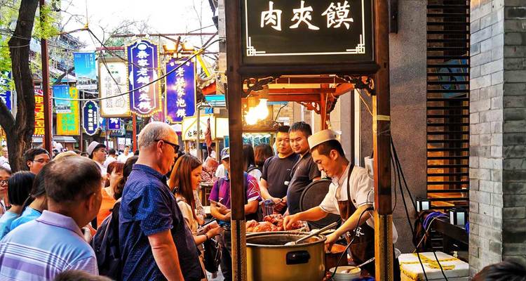 Étal de restauration rapide bondé où un cuisinier prépare des en-cas dans un grand wok tandis que des visiteurs curieux observent.