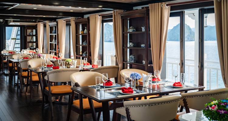 Refined shipboard restaurant with neatly set tables, oceanview windows and decorative shelving.