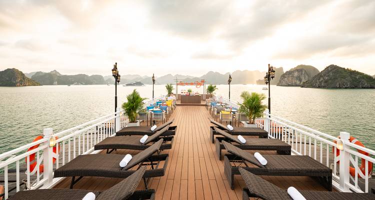 Open-air sun deck lined with loungers on a cruise ship overlooking calm emerald waters and rugged islets at dawn.