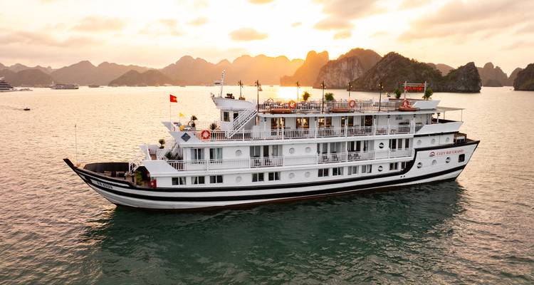 Classic multi-deck cruise vessel sailing Halong Bay at sunset with golden light on distant karst cliffs.