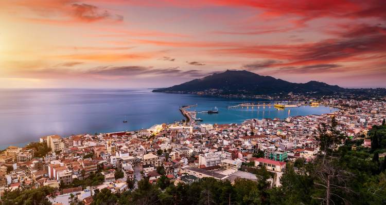 Paisaje urbano al atardecer con un puerto y un fondo montañoso.