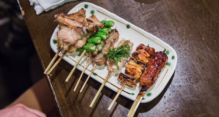 Plato rectangular con brochetas de yakitori variadas colocadas en una mesa de madera en un restaurante japonés.