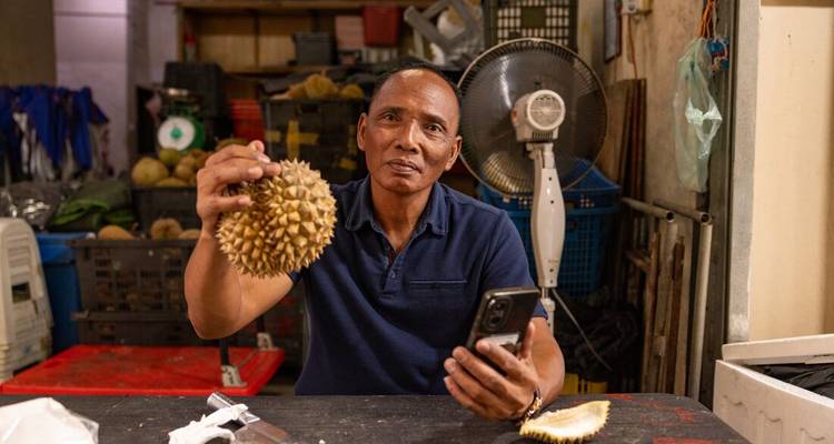 Un homme à l'intérieur d'un étal de marché tient fièrement un durian épineux tout en consultant son téléphone.