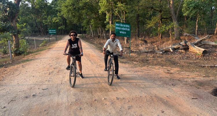 Dos ciclistas andan en bicicleta por un camino de tierra a través del bosque de sal pasando un letrero verde del Pench Tiger Reserve.