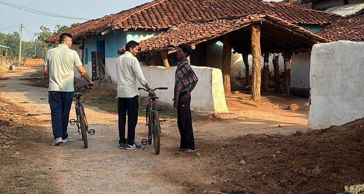 Dos ciclistas charlan con un aldeano fuera de casas rústicas de adobe con techos de teja.