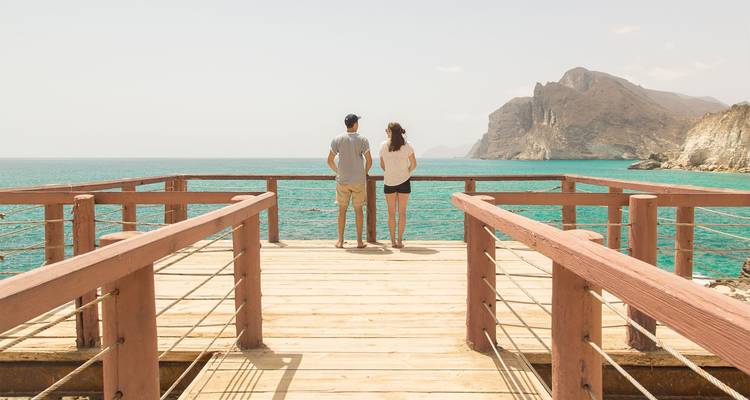 Deux personnes debout sur une terrasse en bois surplombant une mer turquoise.