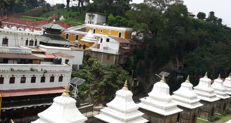Gruppe weißer Schreine und farbenfroher Gebäude auf einem üppigen Flussufer im Kathmandu-Tal.