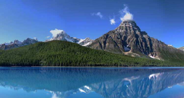 Spiegelglad meer dat perfect een scherpe rotsachtige top en dicht dennenbos onder een felblauwe hemel reflecteert