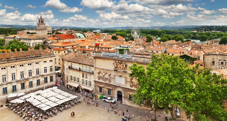 Les toits colorés et la place animée d'Avignon vus de haut sous un ciel de nuages duveteux.