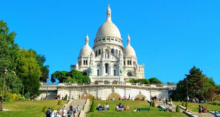 Les dômes blancs de la Basilique du Sacré-Cœur au sommet d'une colline verte remplie de visiteurs sous un ciel bleu éclatant
