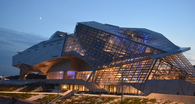 Modern glass-clad museum with angular architecture illuminated at dusk against a fading sky.