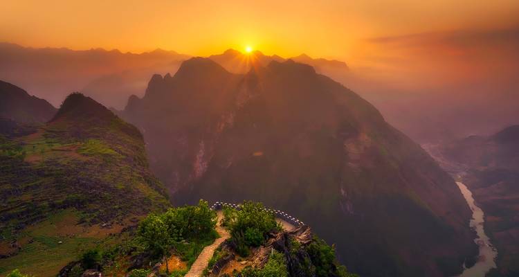 Amanecer dorado sobre crestas montañosas dramáticas y un río sinuoso visto desde un sendero mirador elevado.