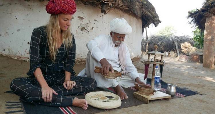 Turista se sienta con un aldeano anciano preparando pan plano tradicional afuera de una cabaña rústica.