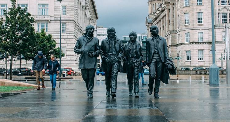 Lebensgroße Beatles-Statuen auf einem nassen Platz mit Fußgängern und historischen Gebäuden in Liverpool.