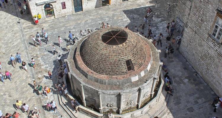 Aerial look at Dubrovnik’s circular Onofrio Fountain amid stone streets and wandering visitors.