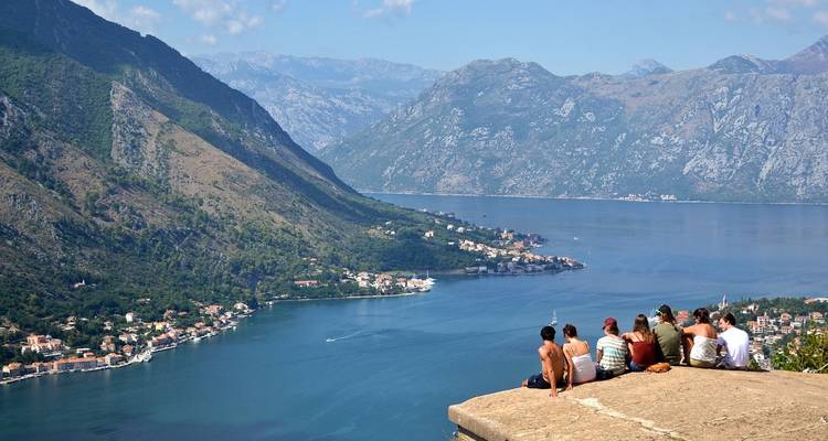 Group of friends sit on a fortress wall overlooking the dramatic bay and mountains of Kotor.