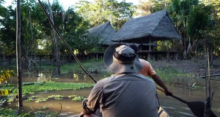 Twee mensen die een boot peddelen richting rieten hutten in een weelderige omgeving.