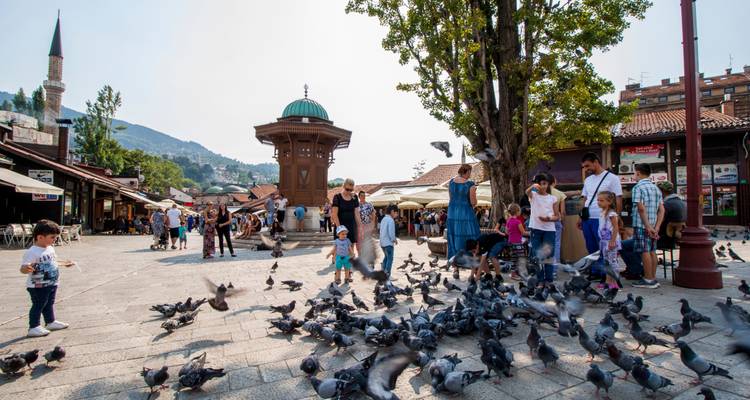 Platz in Sarajevo mit Tauben und Menschen, traditioneller Holzbrunnen in der Mitte.