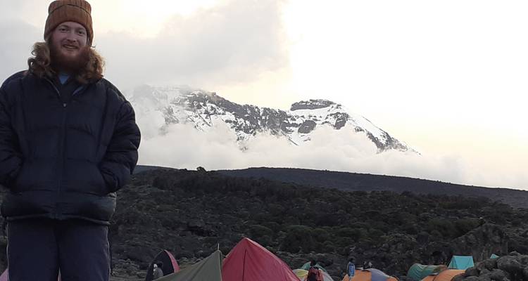 Mann steht neben bunten Zelten auf einem Bergcampingplatz, mit schneebedeckten Gipfeln im Hintergrund.