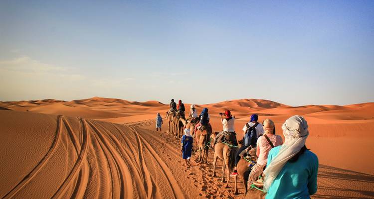 Une caravane de chameaux suit les pistes sableuses à travers les immenses dunes ondulantes sous un ciel bleu pâle.