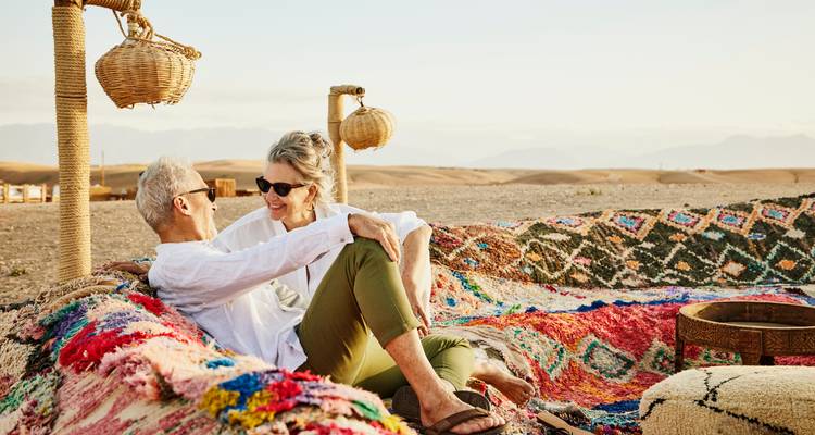 Smiling couple lounging on colorful Berber carpets at a desert camp with distant mountains.