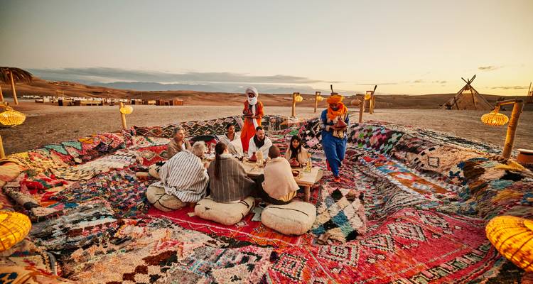 Gruppe versammelt sich zum Abendessen auf Bodenkissen um einen niedrigen Tisch im Wüstenlager bei Sonnenuntergang.