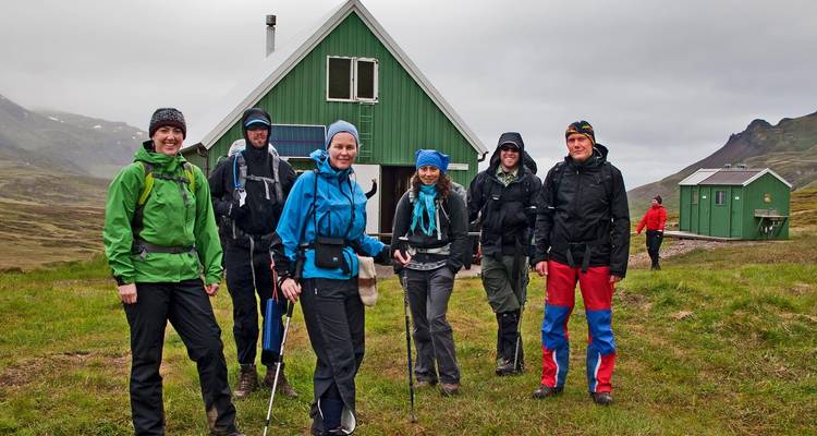 Group of people in hiking gear in front of a cabin in a mountainous area.